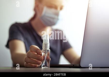 Frau in Maske und Hand mit Alkoholflasche des Desinfektionsmittels in der Nähe des Laptops. Coronavirus covid-19 Prävention am Arbeitsplatz oder zu Hause während der Quarantäne Stockfoto