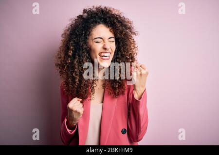 Junge schöne Geschäftsfrau mit lockigen Haaren und Piercing trägt elegante Jacke feiern überrascht und erstaunt für den Erfolg mit erhobenen Armen und Ey Stockfoto
