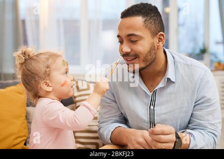 Fröhliches kleines Mädchen mit Spaß malen bunte Striche auf Gesicht ihres glücklichen jungen Vaters Stockfoto