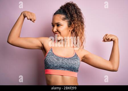 Schöne Sportlerin mit lockigem Haar Sport tragen Sportbekleidung über rosa Hintergrund zeigt Arme Muskeln lächelnd stolz. Fitnesskonzept. Stockfoto