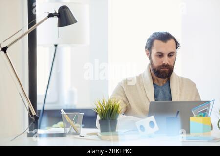 Junge Erwachsene kaukasischen Mann mit Bart auf Gesicht tragen komfortable Outfit sitzen am Schreibtisch arbeiten auf Laptop Stockfoto