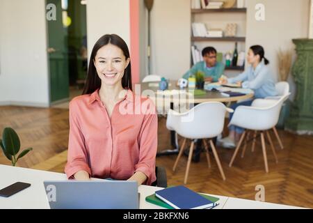 Fröhliche junge Frau in rosa Bluse sitzt am Büroschreibtisch Blick auf Kamera lächelnd, ihre Kollegen hinter ihr arbeiten Stockfoto