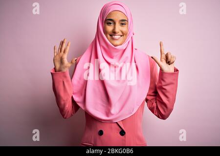 Junge schöne Brünette Geschäftsfrau trägt rosa muslimischen Hijab und Business-Jacke zeigt und zeigt mit den Fingern Nummer sieben während lächelnd c Stockfoto