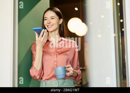 Glückliche junge kaukasische Frau stehend entspannt mit Tasse Kaffee Aufnahme Sprachnachricht auf ihrem Smartphone während der Pause Stockfoto
