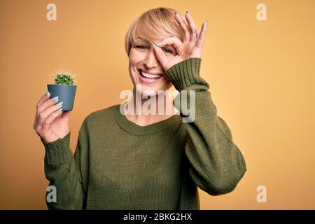 Junge blonde Frau mit kurzen Haaren hält saftigen grünen Kaktus über gelbem Hintergrund mit glücklichen Gesicht lächelnd tun ok Zeichen mit Hand auf Auge Anschauen Stockfoto