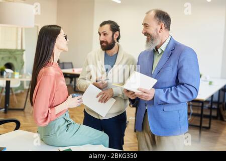 Zwei moderne Männer und junge Geschäftsfrau unterhalten sich während der Kaffeepause bei der Arbeit über etwas Stockfoto