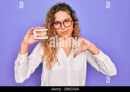 Schöne blonde Frau mit blauen Augen hält Kunststoff Dentrure Zähne über lila Hintergrund mit ärgerlichen Gesicht, negative Zeichen zeigt Abneigung mit Daumen Stockfoto