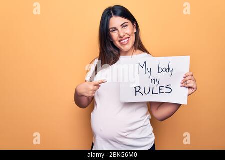 Junge schöne Brünette schwangere Frau mit Banner mit meinem Körper meine Regeln Botschaft lächelnd glücklich zeigen mit Hand und Finger Stockfoto