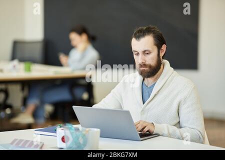 Schöner junger Mann mit Bart auf Gesicht sitzt am Bürotisch arbeiten auf seinem Laptop mit ernsten Gesichtsausdruck Stockfoto
