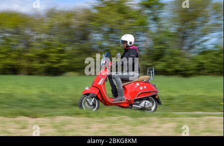 Kiel, Deutschland. Mai 2020. Bei sonnigem Wetter fährt eine Frau mit ihrem roten 125 ccm Vespa Roller auf einer unbefestigten Straße mit Schutzkleidung, Helm und Handschuhen. Seit 01.01.2020 brauchen Inhaber eines Autoführerscheins keinen separaten Motorradführerschein mehr. Nur 5 Fahrstunden und 4 Theoriestunden reichen aus, um Zweiräder bis zu 125 ccm fahren zu dürfen. Quelle: Axel Heimken/dpa/Alamy Live News Stockfoto