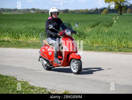 Kiel, Deutschland. Mai 2020. Bei sonnigem Wetter fährt eine Frau mit ihrem roten 125 ccm Vespa Roller auf einer unbefestigten Straße mit Schutzkleidung, Helm und Handschuhen. Seit 01.01.2020 brauchen Inhaber eines Autoführerscheins keinen separaten Motorradführerschein mehr. Nur 5 Fahrstunden und 4 Theoriestunden reichen aus, um Zweiräder bis zu 125 ccm fahren zu dürfen. Quelle: Axel Heimken/dpa/Alamy Live News Stockfoto