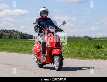 Kiel, Deutschland. Mai 2020. Bei sonnigem Wetter fährt eine Frau mit ihrem roten 125 ccm Vespa Roller auf einer unbefestigten Straße mit Schutzkleidung, Helm und Handschuhen. Seit 01.01.2020 brauchen Inhaber eines Autoführerscheins keinen separaten Motorradführerschein mehr. Nur 5 Fahrstunden und 4 Theoriestunden reichen aus, um Zweiräder bis zu 125 ccm fahren zu dürfen. Quelle: Axel Heimken/dpa/Alamy Live News Stockfoto