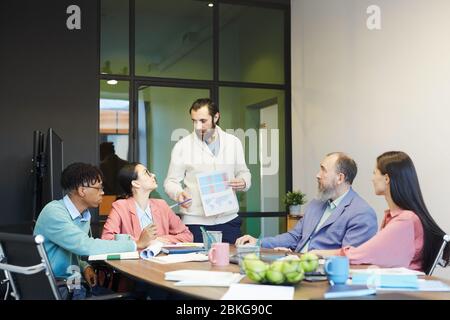 Junger bärtiger Mann, der vor seinen Kollegen im Bürovorstandssaal steht und Papier mit Diagrammen hält, die über Projektproblem sprechen Stockfoto