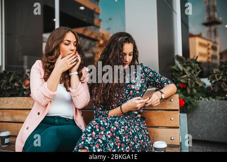 Bild von neugierigen Frau Spionage und guckt auf das Smartphone ihrer Freundin, während sie auf dem Handy reden und auf der Bank in der Stadt Straße sitzen Stockfoto