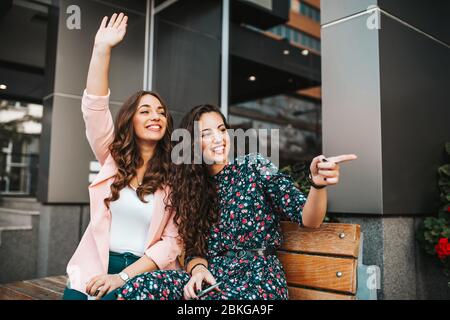 Bild einer fröhlichen, emotionalen jungen, hübschen Freundinnen, die draußen auf der Straße der Stadt sitzen und auf jemanden winken Stockfoto