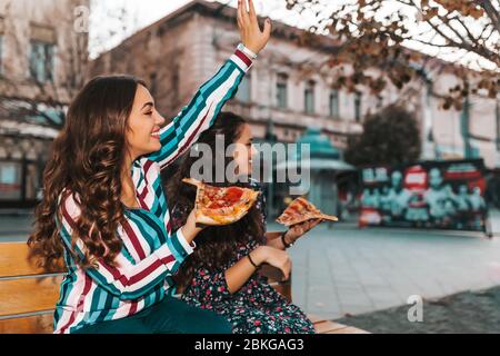 Zwei schöne junge Frauen, die im Freien Pizza essen und ihre Freunde winken. Lifestyle, modernes Leben. Stockfoto