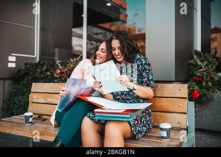 Glückliche Freundinnen, Studenten lesen zusammen, gute Nachrichten, während sie draußen sitzen. Zwei fröhliche Frauen feiern Erfolg. Stockfoto