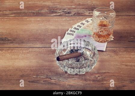 Ein Glas Whisky und ein Geld mit kubanischer Zigarre auf einem braunen Holztisch. Draufsicht. Stockfoto