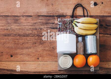 Papiertüte mit Lebensmitteln, Toilettenpapier und Handdesinfektionsmittel darauf. Flach auf dem Holztisch liegen. Flach auf dem Holztisch liegen. Covid-19 Ausbruch, Selbstisolierung, Lieferung, Spendenkonzept. Stockfoto
