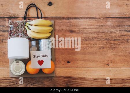 Papiertüte mit Lebensmitteln, Toilettenpapier, Handdesinfektionsmittel und Karte mit Schild Stay Safe darauf. Flach auf dem Holztisch liegen. Covid-19 Ausbruch, Selbstisolierung, Lieferung, Spendenkonzept. Stockfoto