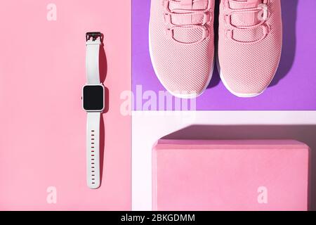 Neue lila Sneaker, rosa Yoga-Block und Smart-Uhr auf buntem Hintergrund. Overhead-Aufnahme von Laufschuhen. Draufsicht, flaches Lay Stockfoto