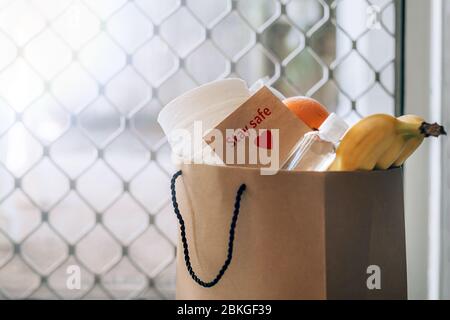 Papiertüte mit Lebensmitteln, Toilettenpapier, Handdesinfektionsmittel und Karte mit Schild Bleib sicher auf dem Boden am Eingang zum Haus. Covid-19 Ausbruch, Selbstisolierung, Lieferung, Spende. Stockfoto