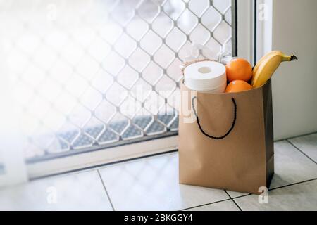 Papiertüte mit Essen und Toilettenpapier auf dem Boden am Eingang zum Haus. Covid-19 Ausbruch, Selbstisolierung, Lieferung, Spende. Stockfoto