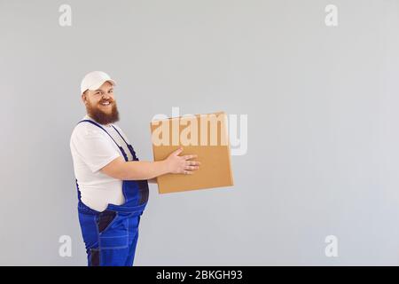 Lustige fette bärtige Lieferung Kurier Lader Mann in einer blauen Uniform mit einem Karton auf einem grauen Hintergrund. Stockfoto