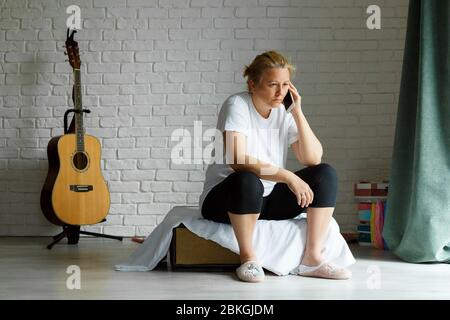 Traurige Frau, die im Wohnzimmer am Telefon telefoniert und im Bus sitzt. 40 Jahre Dame in Hauskleidung. Stockfoto