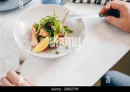 Salat mit Garnelen, Rucola, Zitrone, Tomaten in tiefer Schüssel, mit Händen von Männern, hält Besteck Gabel. Heller Hintergrund. Draufsicht. Stockfoto