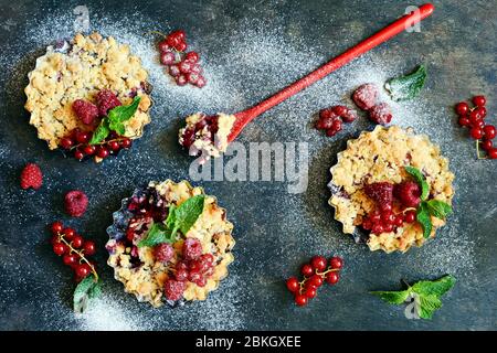 Portionsbeere zerbröckelt in einer Backform auf dunklem Hintergrund. Beerentartlets mit Mürbeteig und saftiger Füllung. Leichte Sommerdessert. Stockfoto