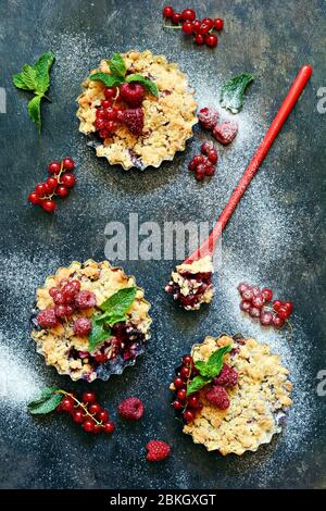 Portionsbeere zerbröckelt in einer Backform auf dunklem Hintergrund. Beerentartlets mit Mürbeteig und saftiger Füllung. Leichte Sommerdessert. Stockfoto