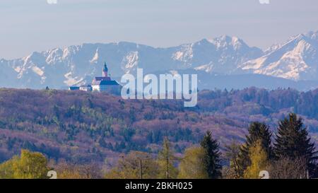 Blick auf Kloster Andechs. Im Hintergrund die schneebedeckte Wettersteinkette. Im Vordergrund Wald. Panoramafarma. Stockfoto