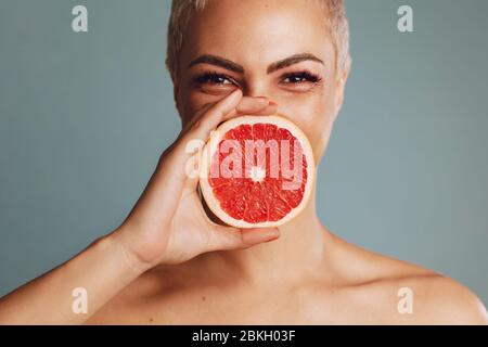 Nahaufnahme einer Frau, die vor grauem Hintergrund eine Grapefruit vor ihrem Mund hält. Schönes weibliches Modell mit einer Grapefruit-Scheibe. Stockfoto