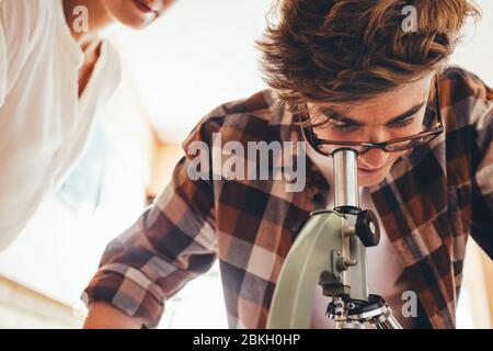 Junge mit Lehrerin im Schullabor, die in ein Mikroskop schaut. Student, der Objektträger durch ein Mikroskop im Unterricht betrachtet. Stockfoto