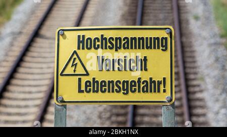 Gelbes Schild mit der Aufschrift 'Hochspannung, Sicht, Lebensgefahr'. Quadratisches Format. Im Hintergrund Eisenbahnschienen Stockfoto