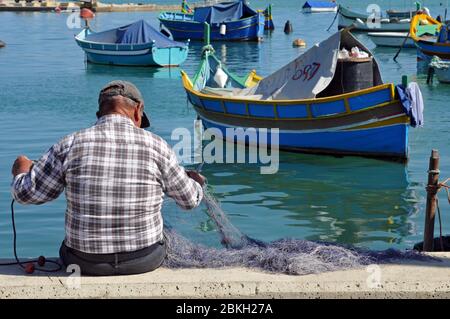 Ein Mann sitzt am Wasser und repariert ein Fischernetz im Dorf Marsaxlokk, Malta. Traditionelle maltesische Boote sind vor der Küste verankert. Stockfoto