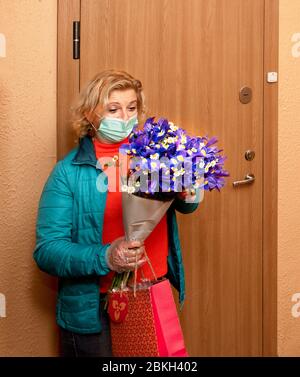 Kurier in Schutzmaske und in medizinischen Handschuhen liefert Blumen und Geschenk an Seniorkunden zum Geburtstag. Lieferservice unter Quarantäne,Krankheit o Stockfoto