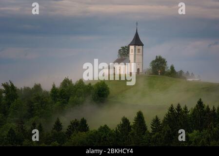 Jamnik Kirche auf einem Hügel im Frühling, neblig Wetter bei Sonnenuntergang in Slowenien, Europa. Berglandschaft kurz nach dem Frühlingsregen. Slowenische Alpen. Stockfoto