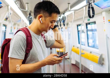 Nahaufnahme eines jungen Mannes, der Musik mit kabellosen Ohrhörern und Mobiltelefonen während der Fahrt mit dem Zug hört. Asian Kerl genießen Musik auf dem Go. Stockfoto