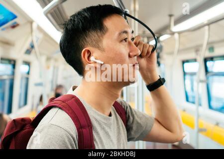 Nahaufnahme des jungen Mannes, der Musik mit kabellosen Ohrstöpseln hört, während er mit dem Zug pendelt. Asiatischer Kerl, der unterwegs Musik genießt. Stockfoto