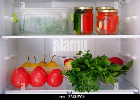 Offener Kühlschrank. Gesunde Rohkost: Birnen, Petersilie, Gurken, Tomaten, Pfeffer. Einmachungen im Glas und Gemüse auf dem Regal des Kühlschranks. Stockfoto