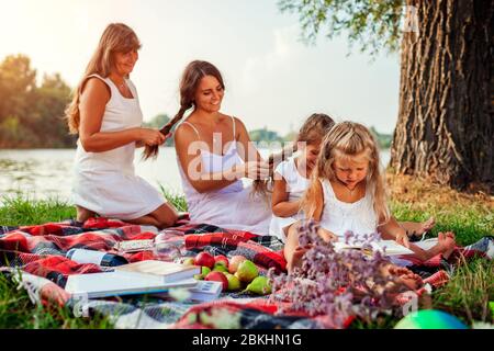 Muttertag. Mutter, Großmutter und Kinder, die Haarzöpfe miteinander webten. Familie, die Spaß beim Picknick im Park hat. Stockfoto