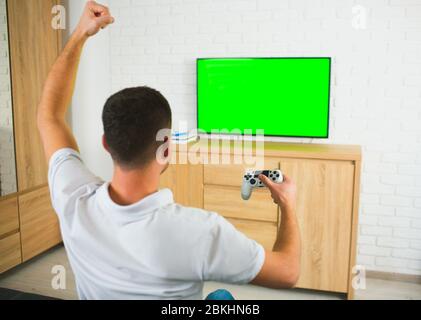 Junger Mann sitzt während der Quarantäne im Zimmer. Guy Computerspiele mit Joystick und tv. Grüner Bildschirm. Alleine spielen und jubeln. Rückansicht. Stockfoto