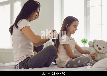 Mutter macht Frisur zu Mädchen Kind auf dem Bett im Schlafzimmer sitzen. Stockfoto