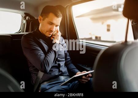 Ein geschäftiger Geschäftsmann im Taxi. Multitasking-Konzept. Der Passagier fährt auf dem Rücksitz und arbeitet gleichzeitig. Spricht auf dem Smartphone und verwendet Tablet-pc. Stockfoto