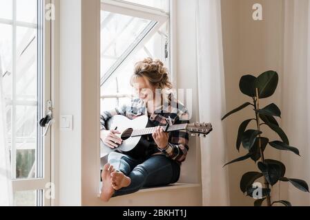 Frau saß zu Hause auf einer Fensterleiste lächelnd spielend Gitarre Stockfoto