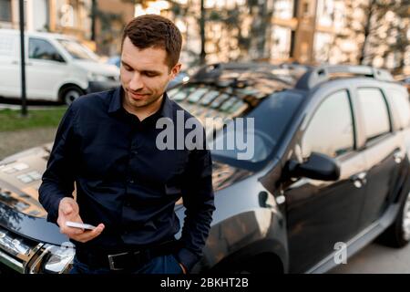 Ein junger Mann oder Geschäftsmann in einem dunklen Hemd steht auf der Straße in der Nähe des Autos wird auf das Smartphone schauen. Der Fahrer wartet auf seinen Passagier oder Stockfoto