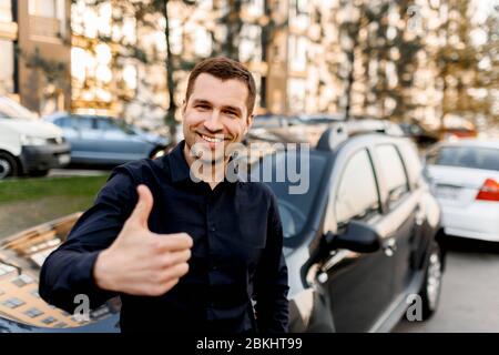 Ein junger Mann steht neben einem Auto und zeigt einen Daumen nach oben.Taxifahrer schaut auf die Kamera und lächelt. Städtische Umwelt und Verkehr Stockfoto