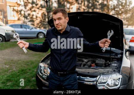 Konzept für Pannengefahren. Das Auto startet nicht. Der junge Mann versucht alles selbst zu reparieren, aber er weiß nicht, was er tun soll. Sie können das Auto nicht reparieren Stockfoto